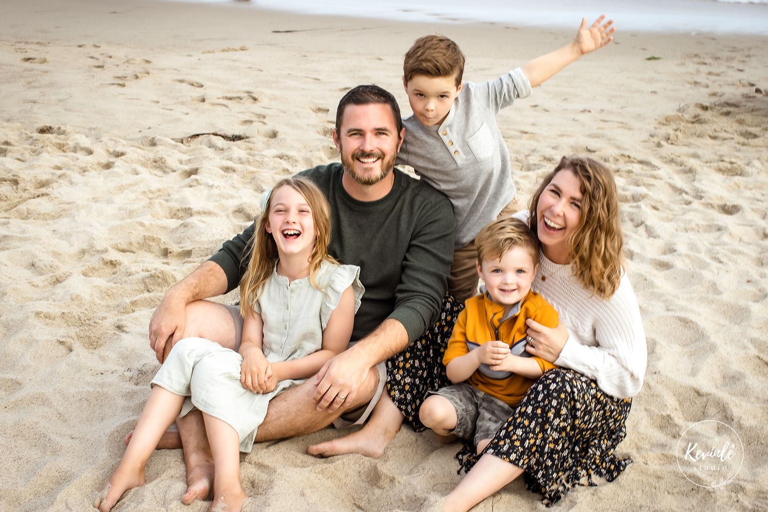 Happy family of five sitting on sandy beach, parents smiling with three young children, ocean backdrop.