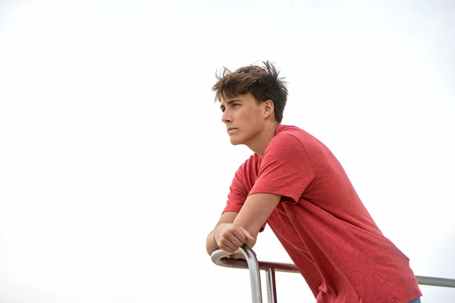 A teenage boy in a red t-shirt leans on a metal railing, gazing thoughtfully into the distance.