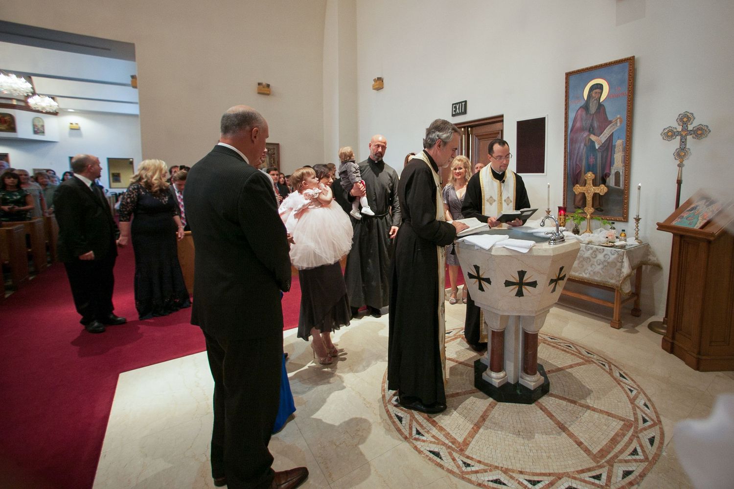 Orthodox baptism ceremony with priest and guests gathered around a baptismal font in a decorated church.