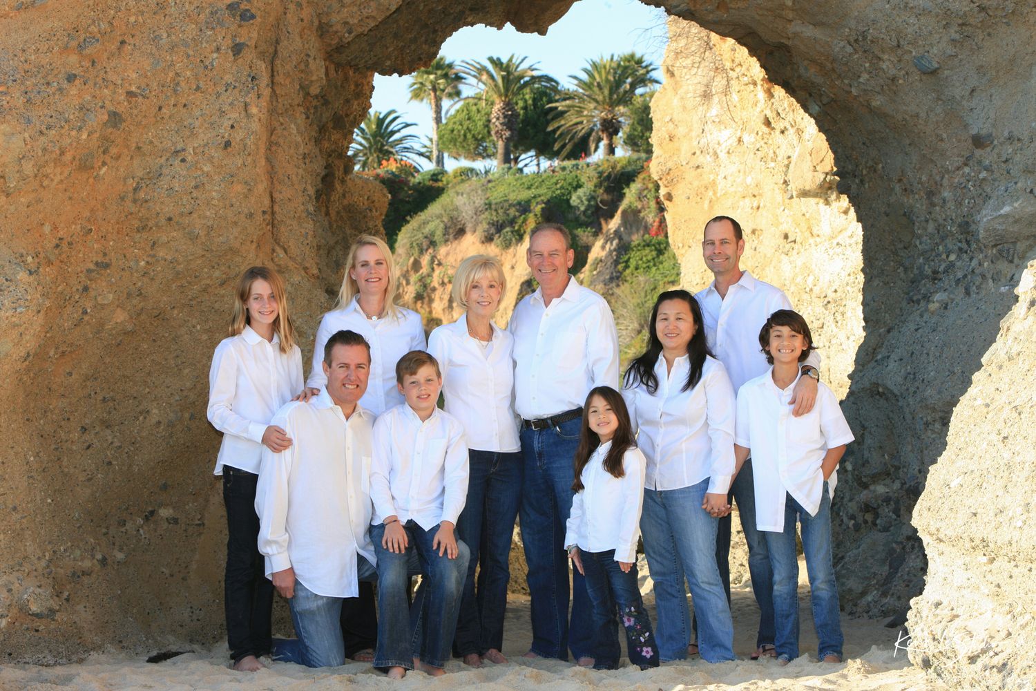Extended family of ten posing together inside a natural rock arch on beach.