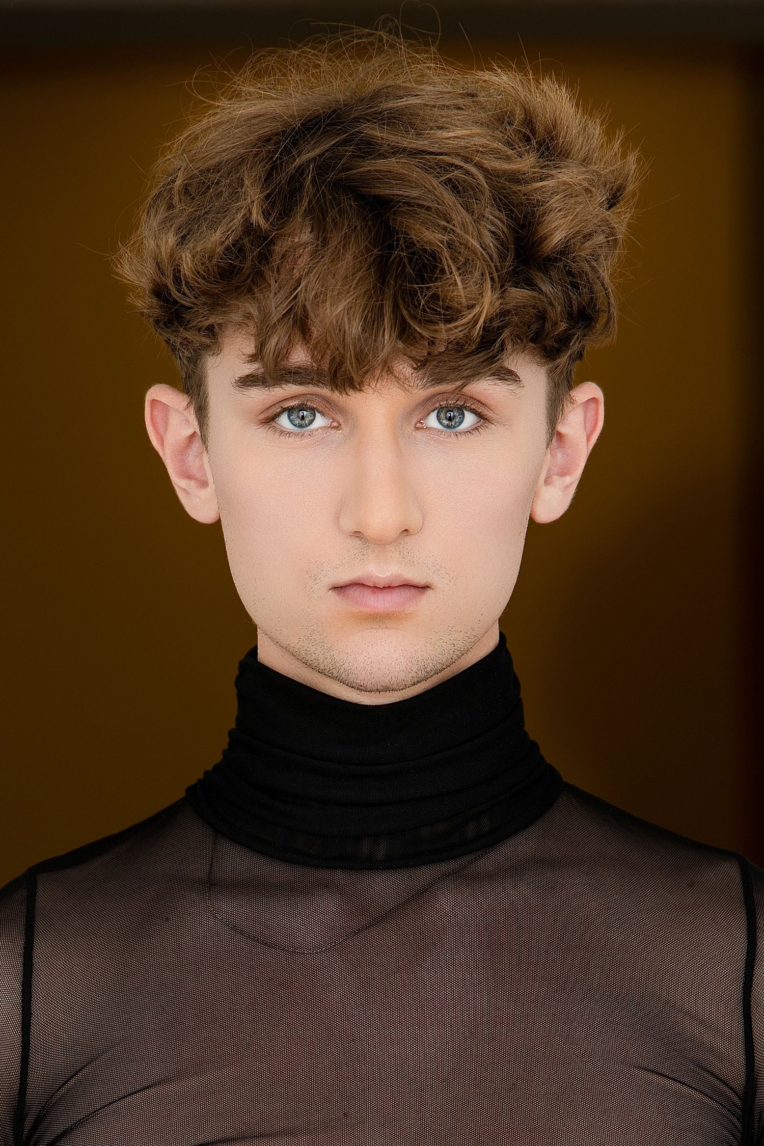 Young man with curly brown hair and blue eyes wearing black turtleneck mesh top, studio portrait.