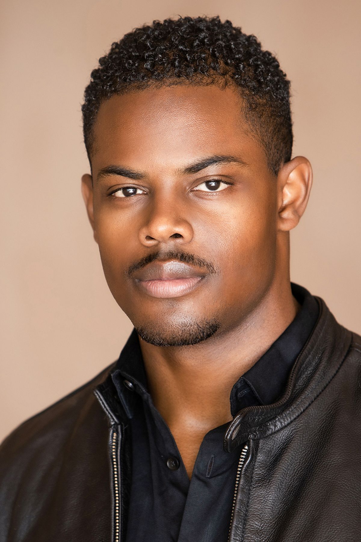 Professional headshot of a young Black man with short curly hair wearing a black leather jacket.