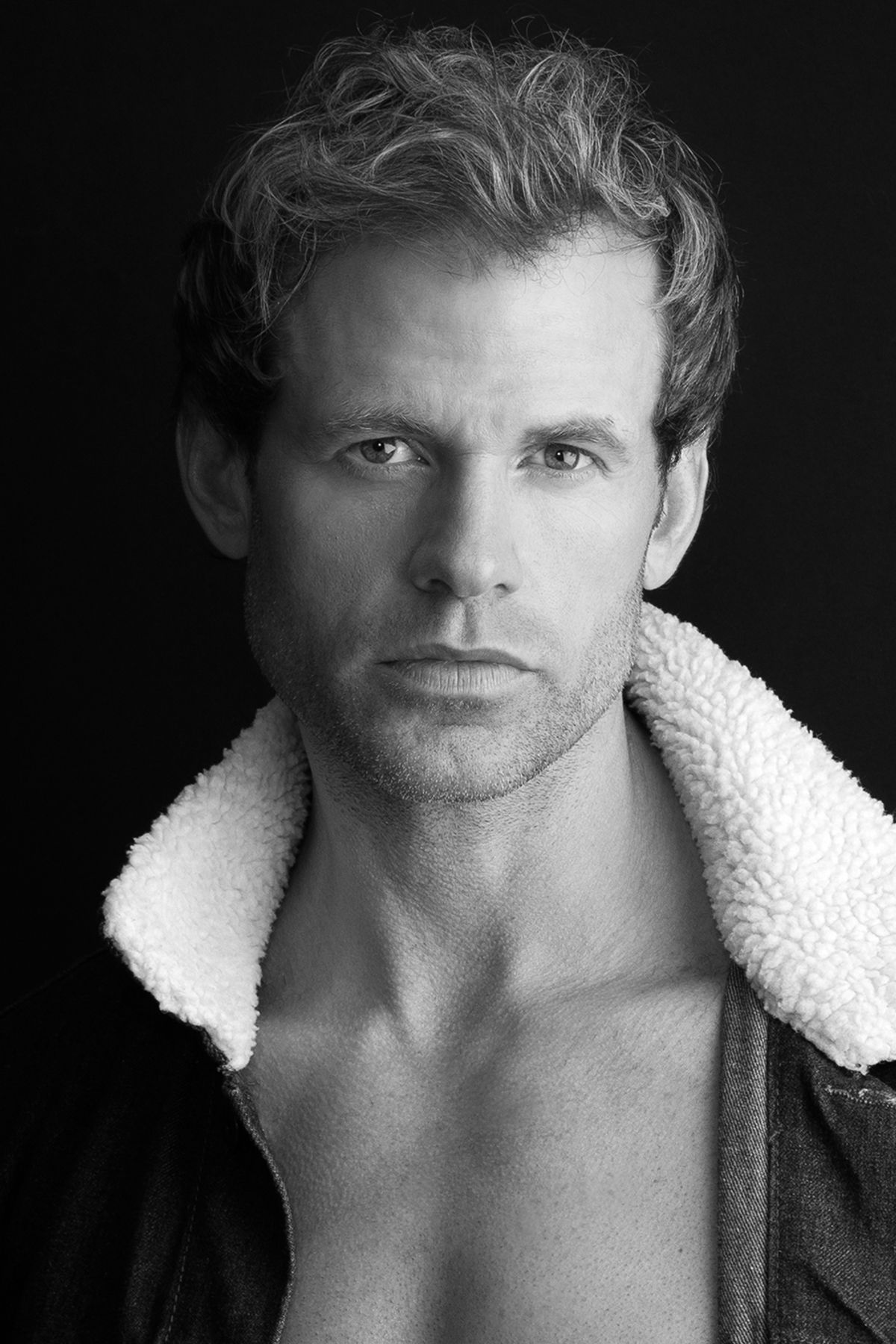 Black and white portrait of a muscular blond man wearing an open sherpa-lined jacket.