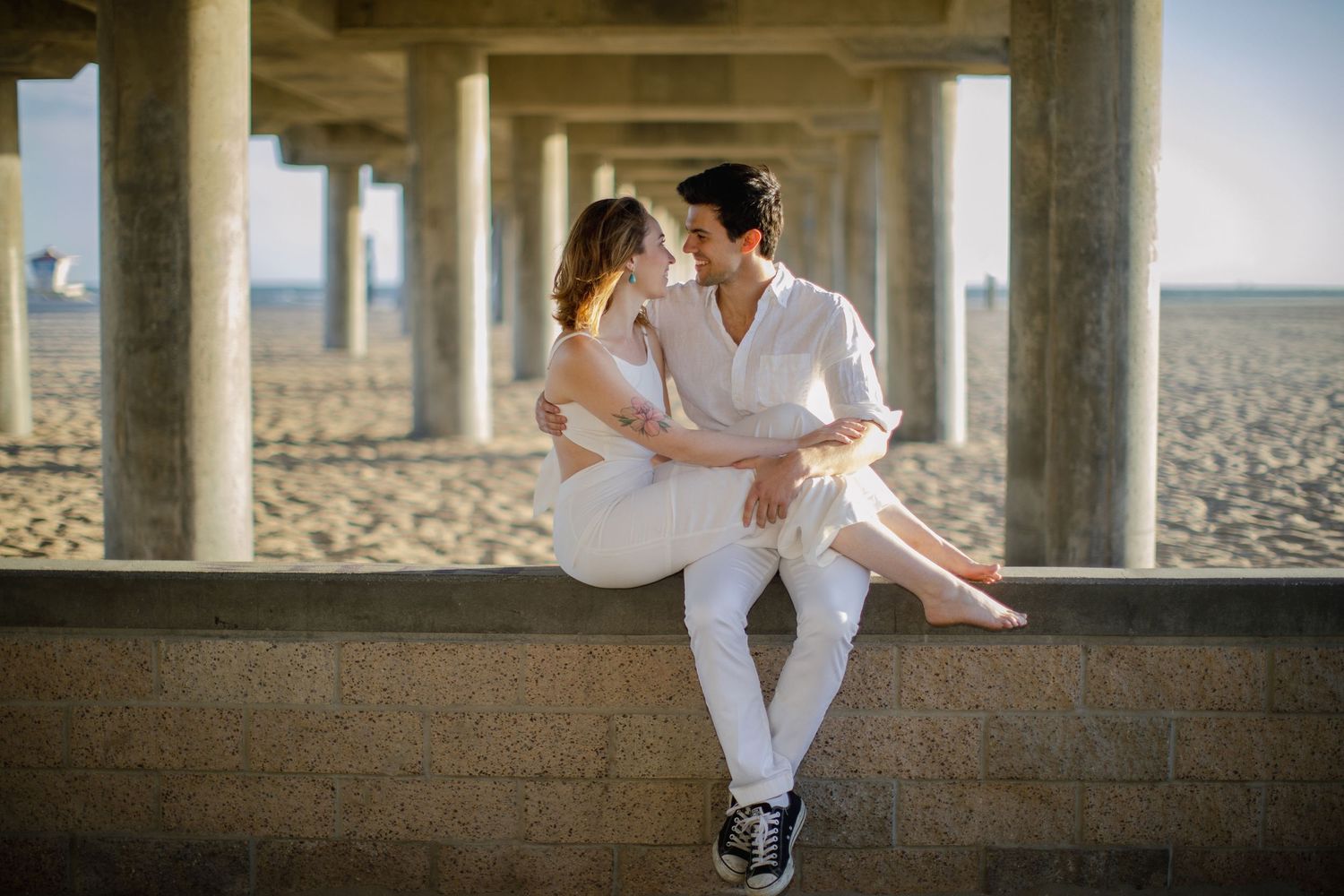 Romantic couple in white outfits share intimate moment sitting on concrete wall at sandy beach.