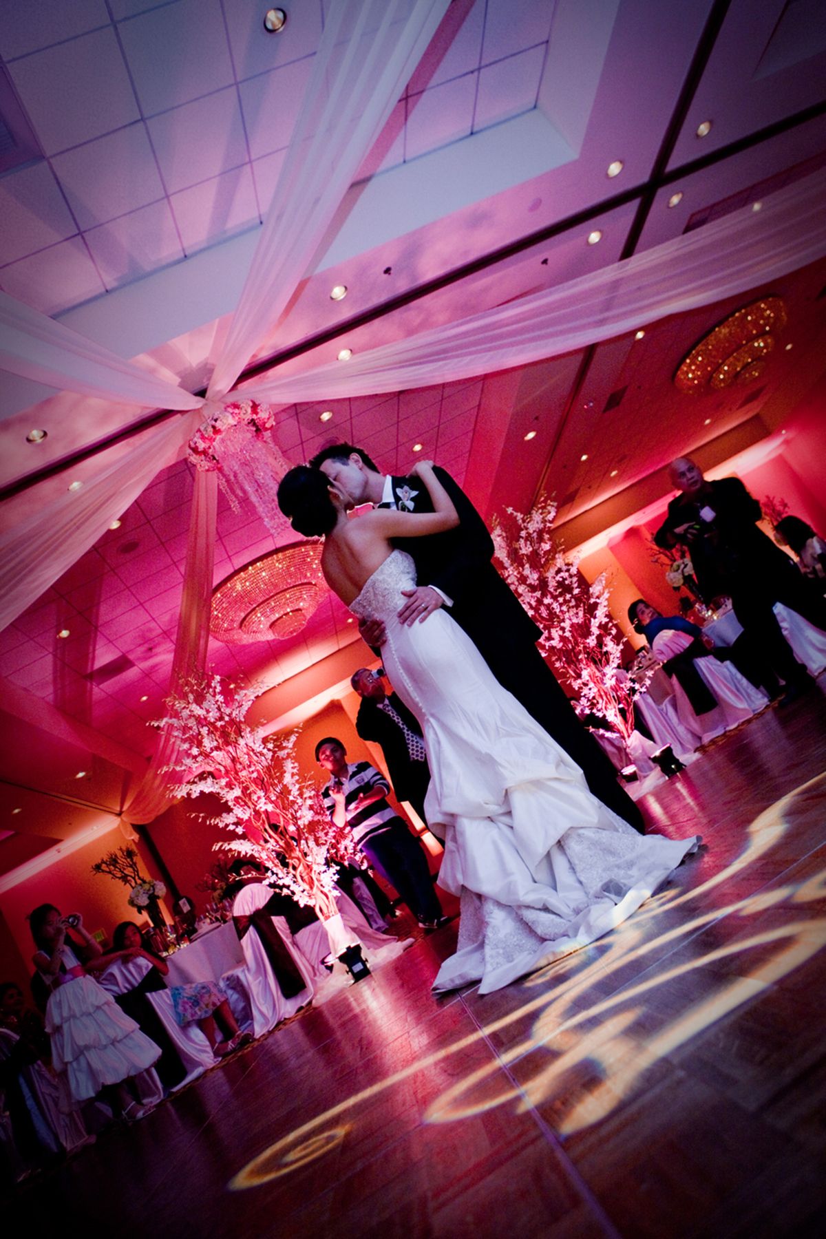 Bride and groom share first kiss on dance floor at pink-lit wedding reception.