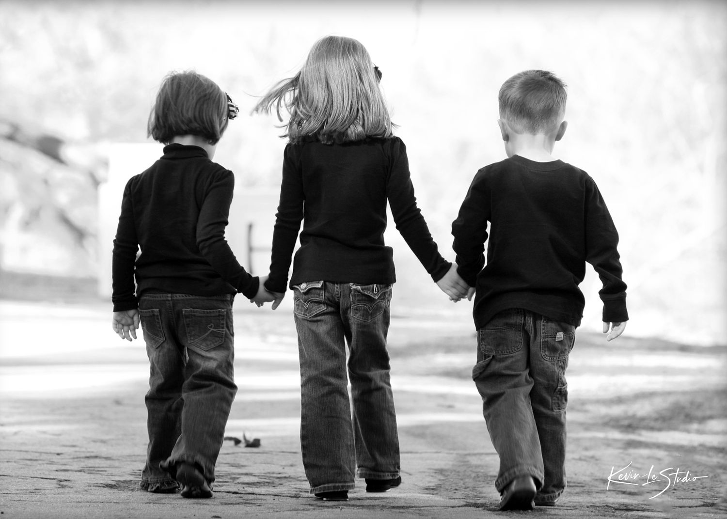 Three children holding hands walking away in a black and white photo, dressed in dark clothing.