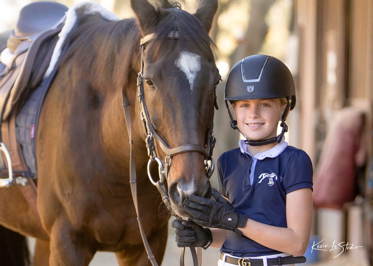 Young equestrian girl in helmet and gloves stands beside a brown horse at a stable, smiling proudly.
