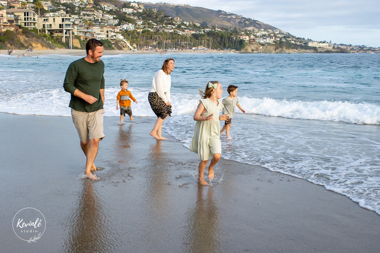 Family laughing and running along a sunny California beach with ocean waves and hillside homes in the background.