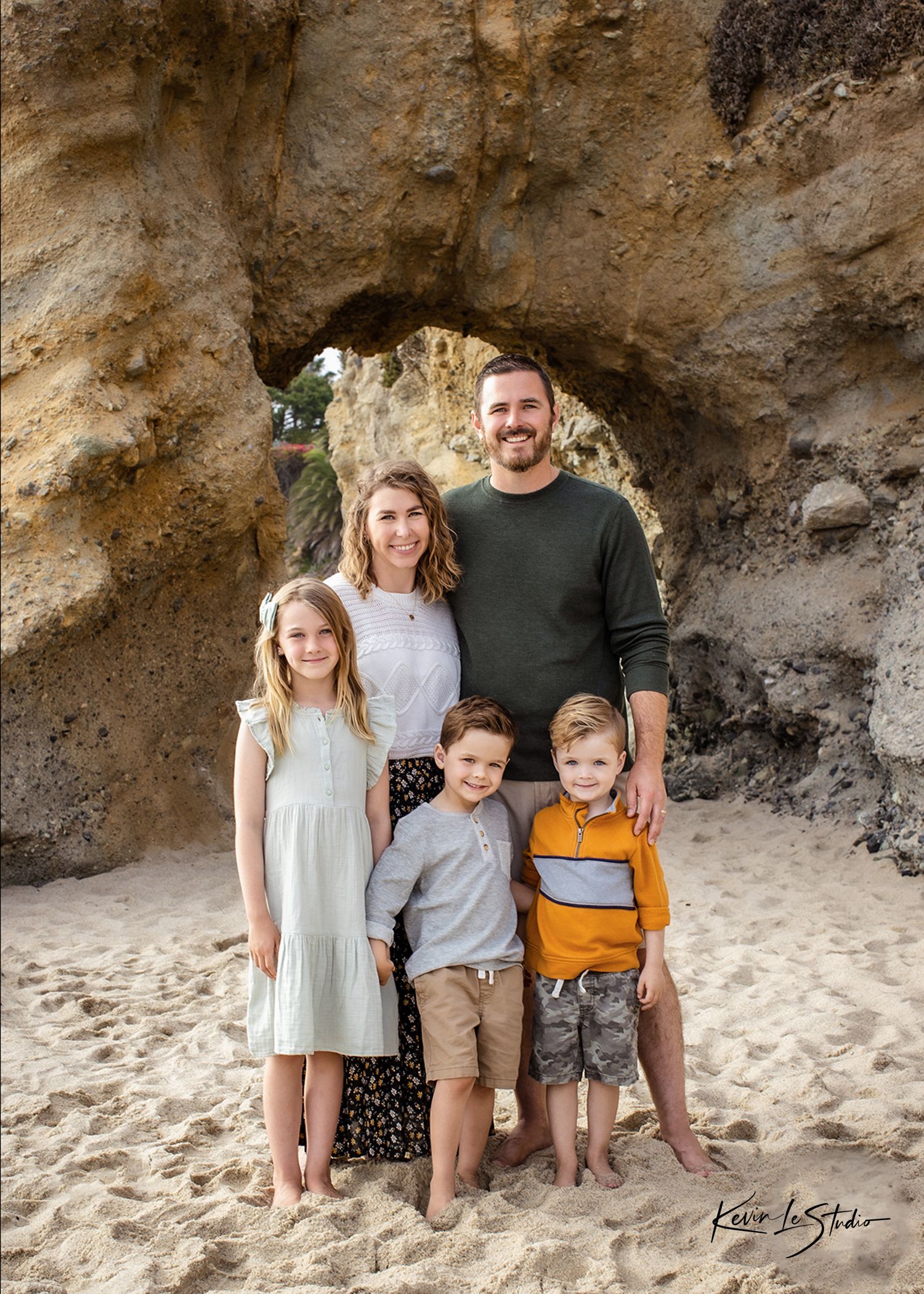 Family of five posing on sandy beach near rocky arch, parents with three young children smiling outdoors.