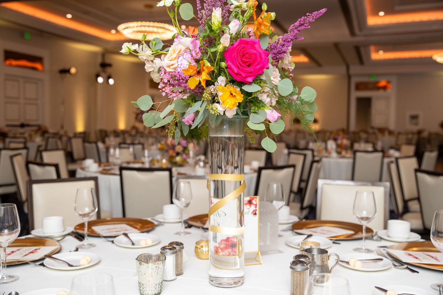 Elegant wedding reception centerpiece with vibrant pink and orange flowers in tall glass vase on white tablecloth.