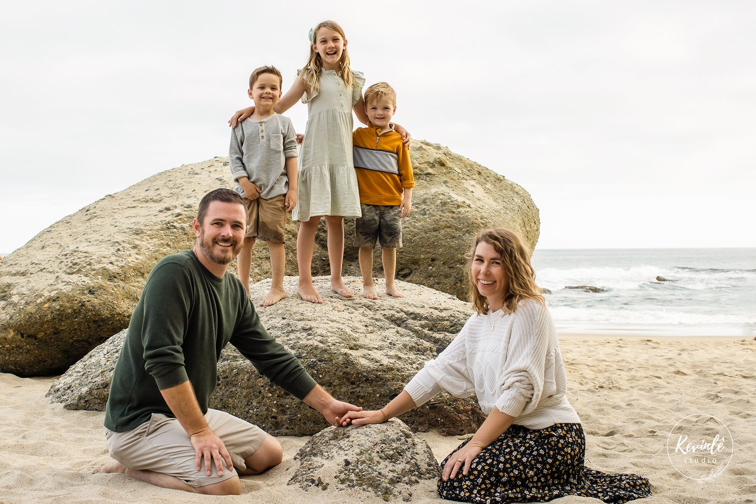 Family of five posing on a sandy beach, parents sitting while three children stand on a large rock with ocean waves behind them.
