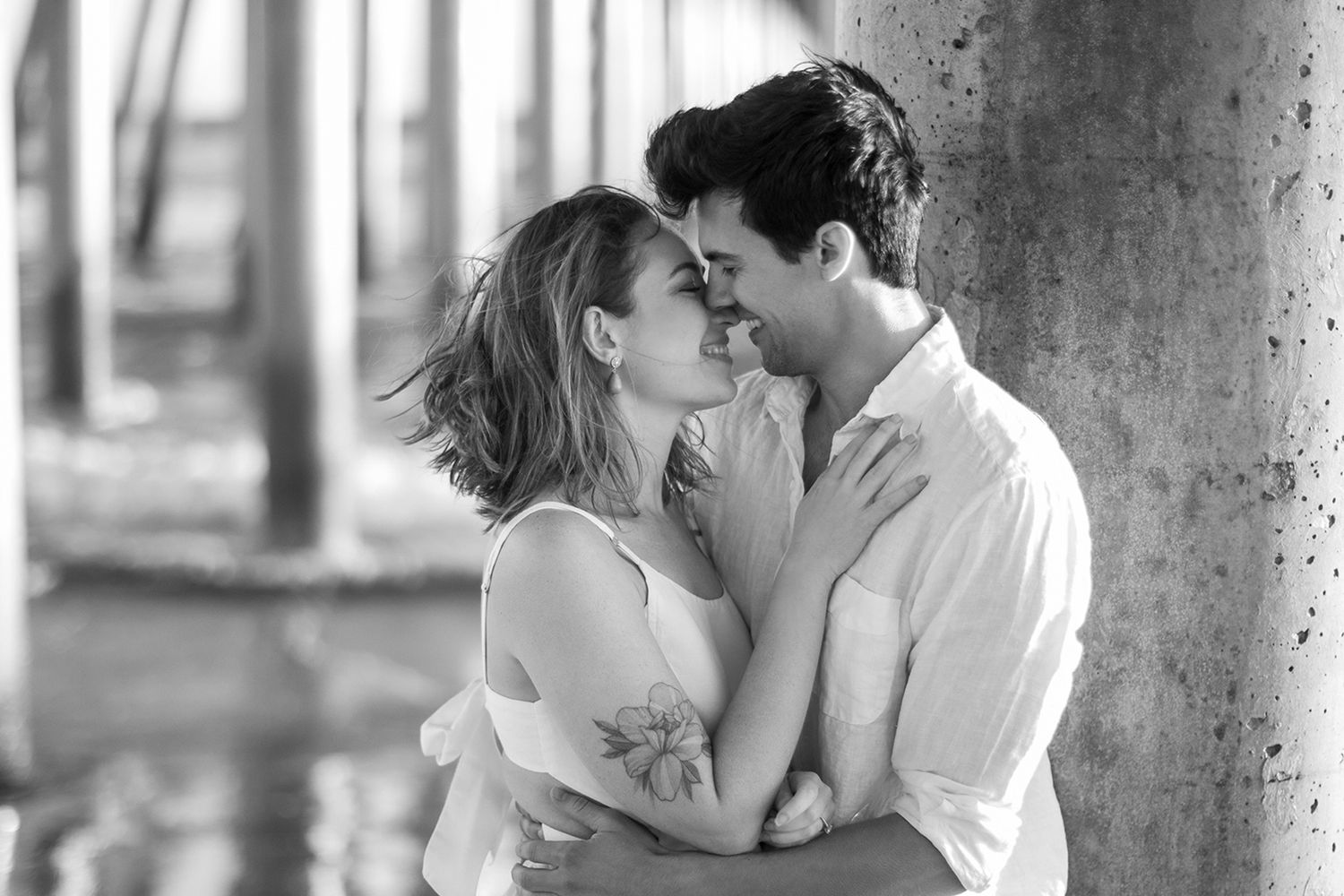 Black and white photo of a romantic couple embracing and gazing at each other under a pier near water.