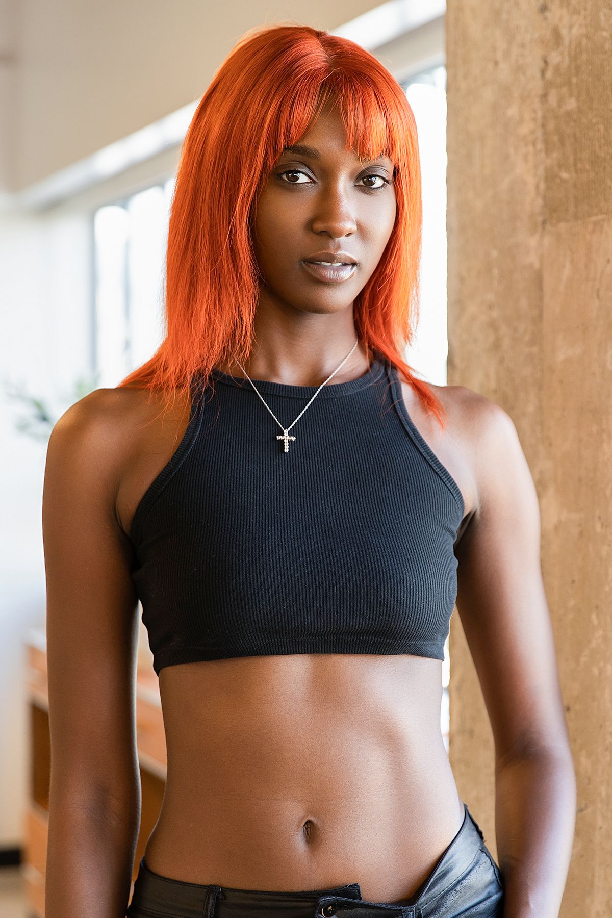 Tall Black woman with bright orange straight wig wearing a black crop top and jeans with a cross necklace.