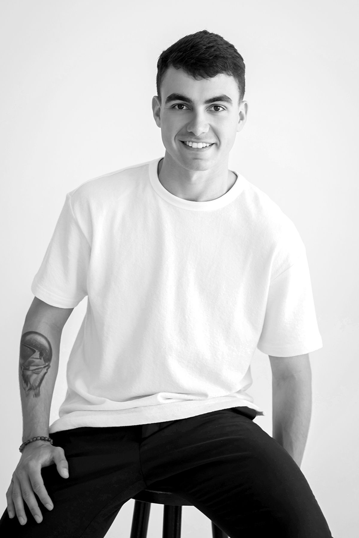 Black and white photo of a smiling young man in a white t-shirt sitting on a stool with a tattoo.