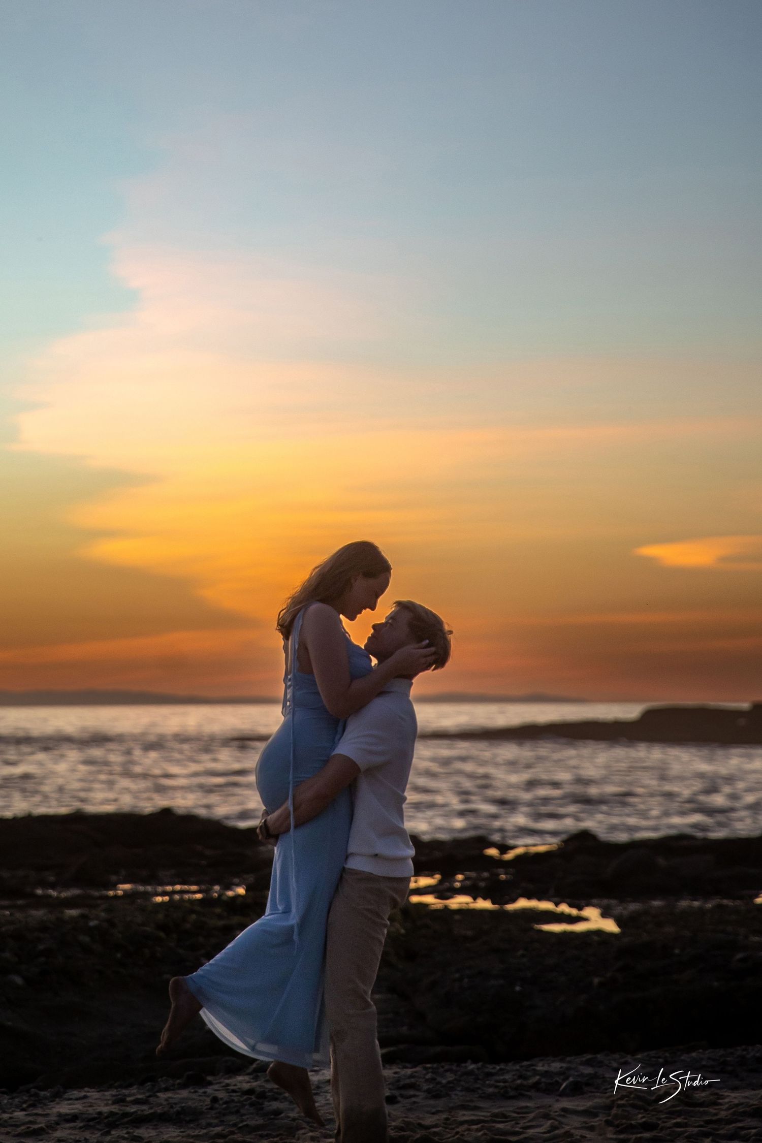 Romantic couple embracing at sunset on a rocky beach, woman lifted in mans arms with vibrant orange sky behind them.
