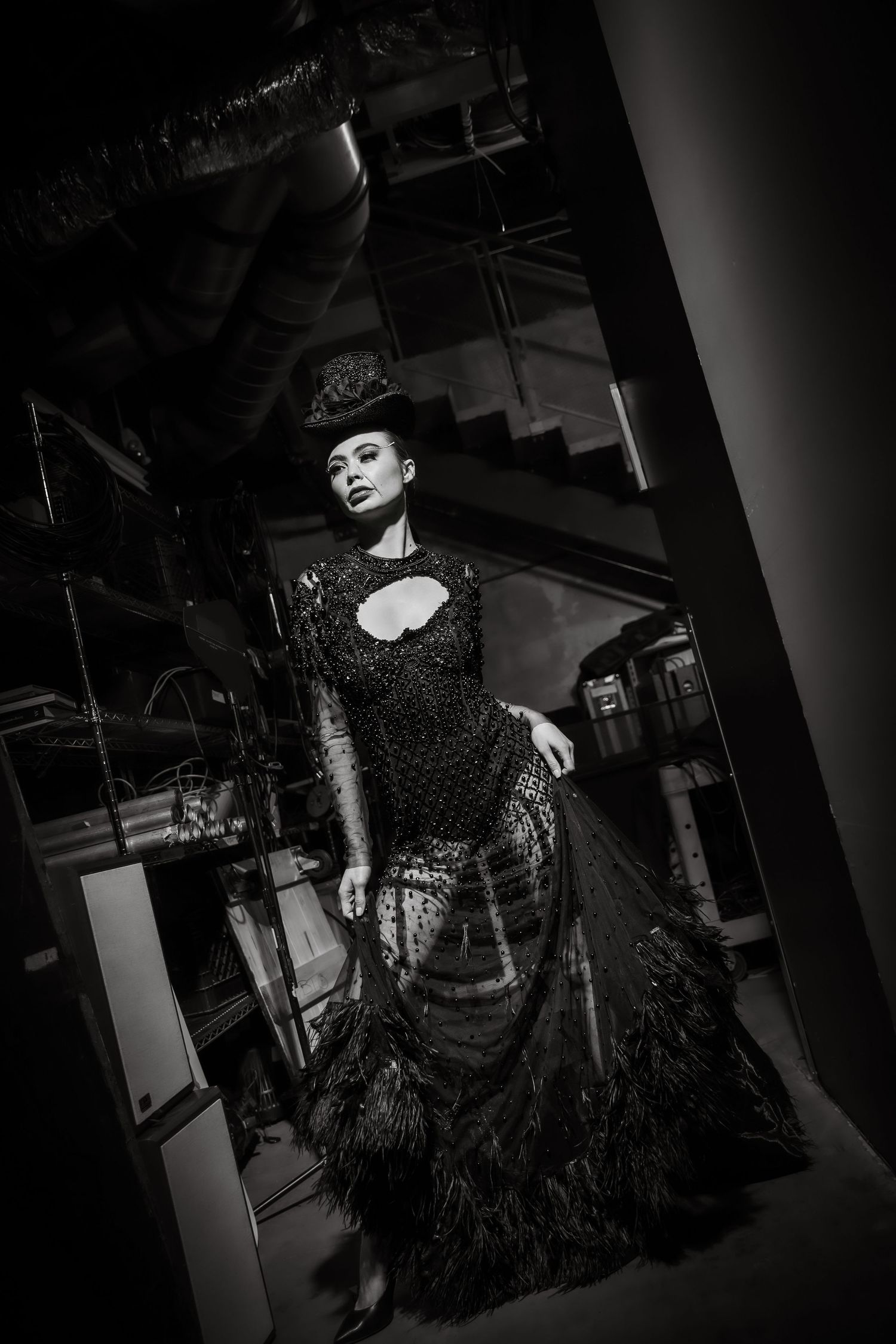 Black and white fashion photo of a model in an ornate black feathered gown and hat in an industrial setting.