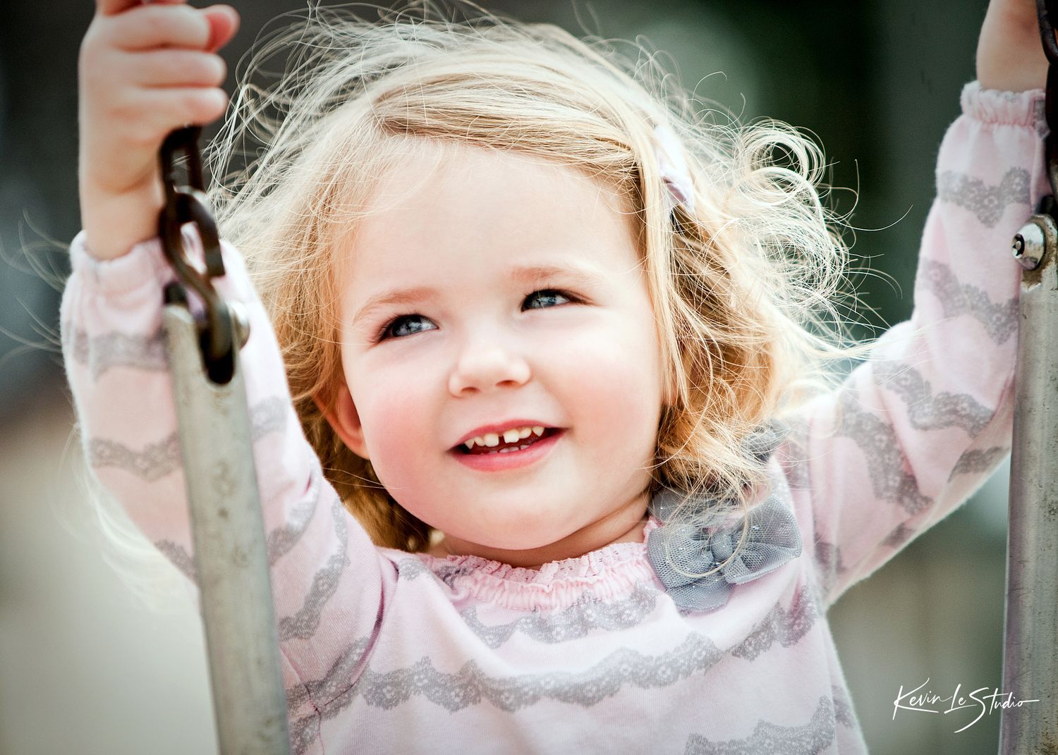 Smiling toddler girl with blonde hair holding swing chains, wearing pink floral top, outdoors in natural light.