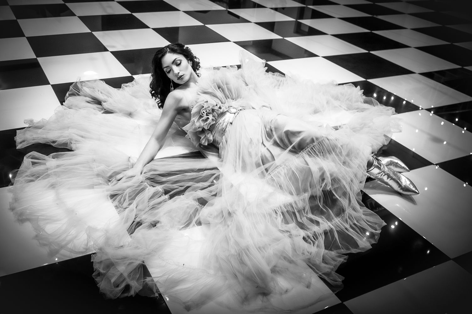 Elegant bride in a flowing tulle gown lying on a checkered floor in a stunning black and white photo.