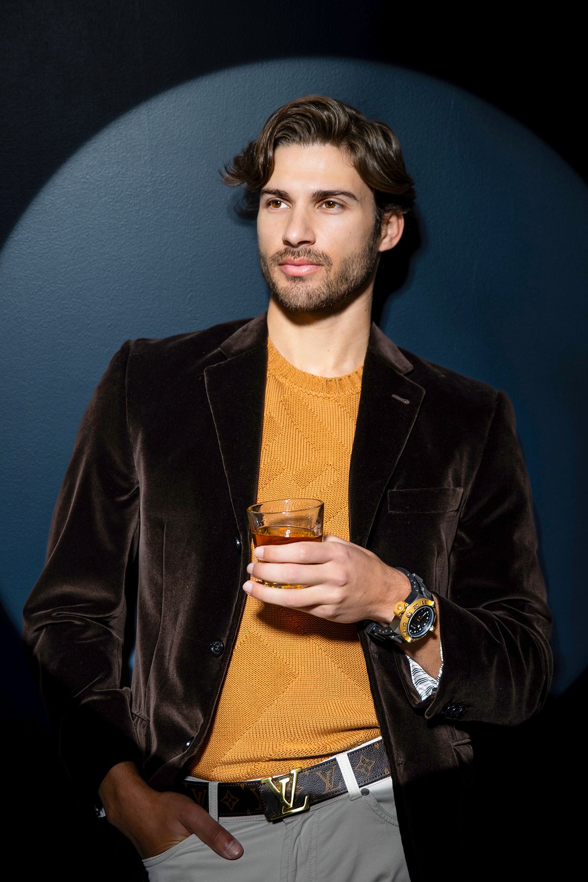 Stylish man in brown velvet blazer and orange sweater holding whiskey glass with luxury watch and Louis Vuitton belt.