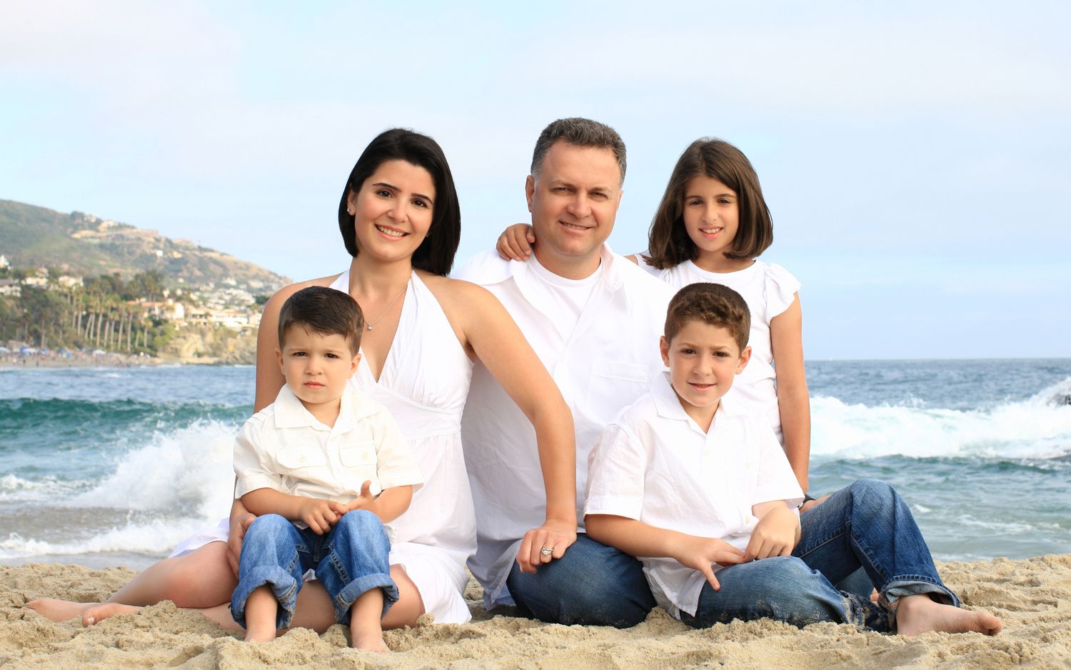 Family of five in white outfits posing on a sunny beach with ocean waves in the background.