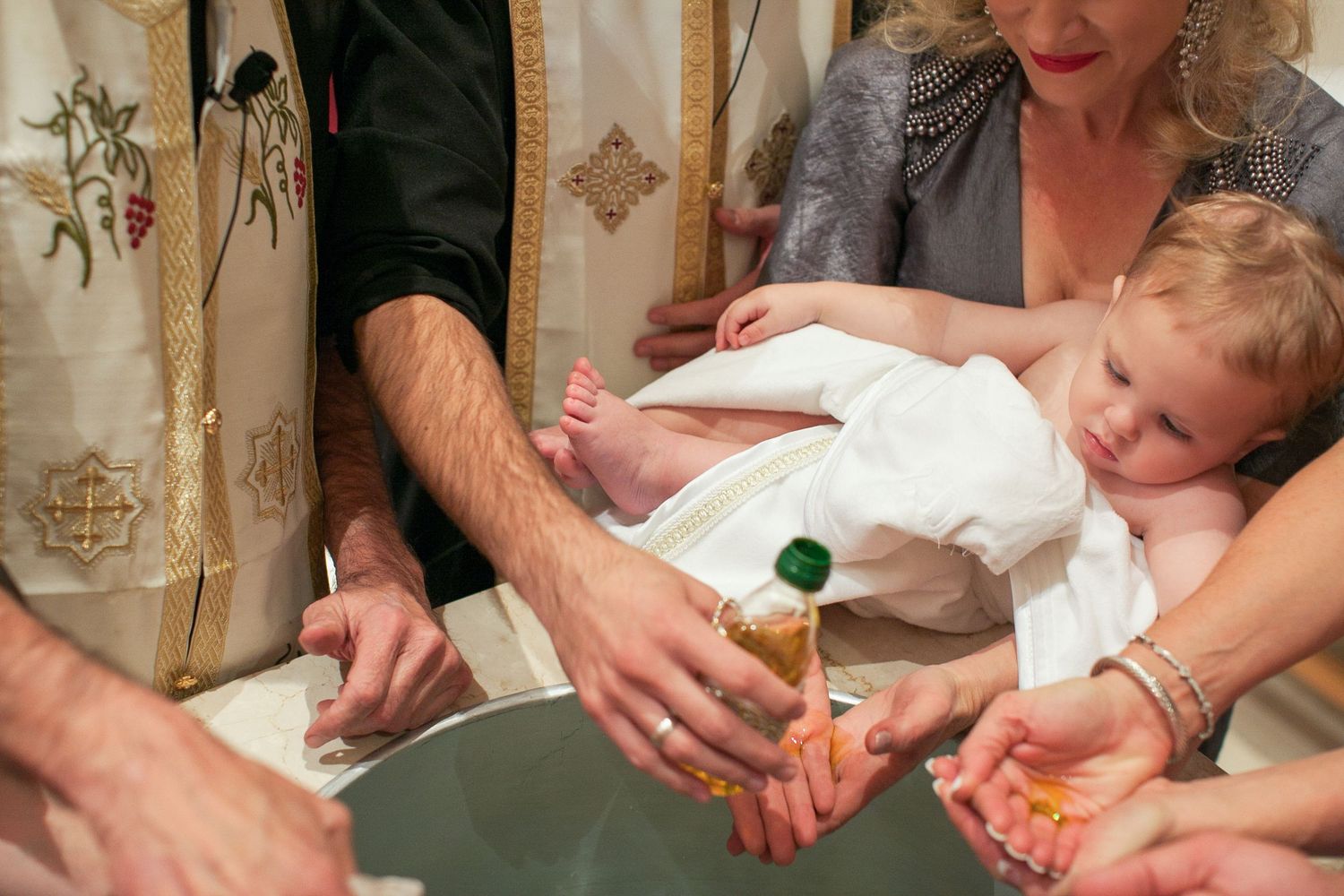 Baby in white gown being held during a Christian baptism ceremony with holy water and oil.