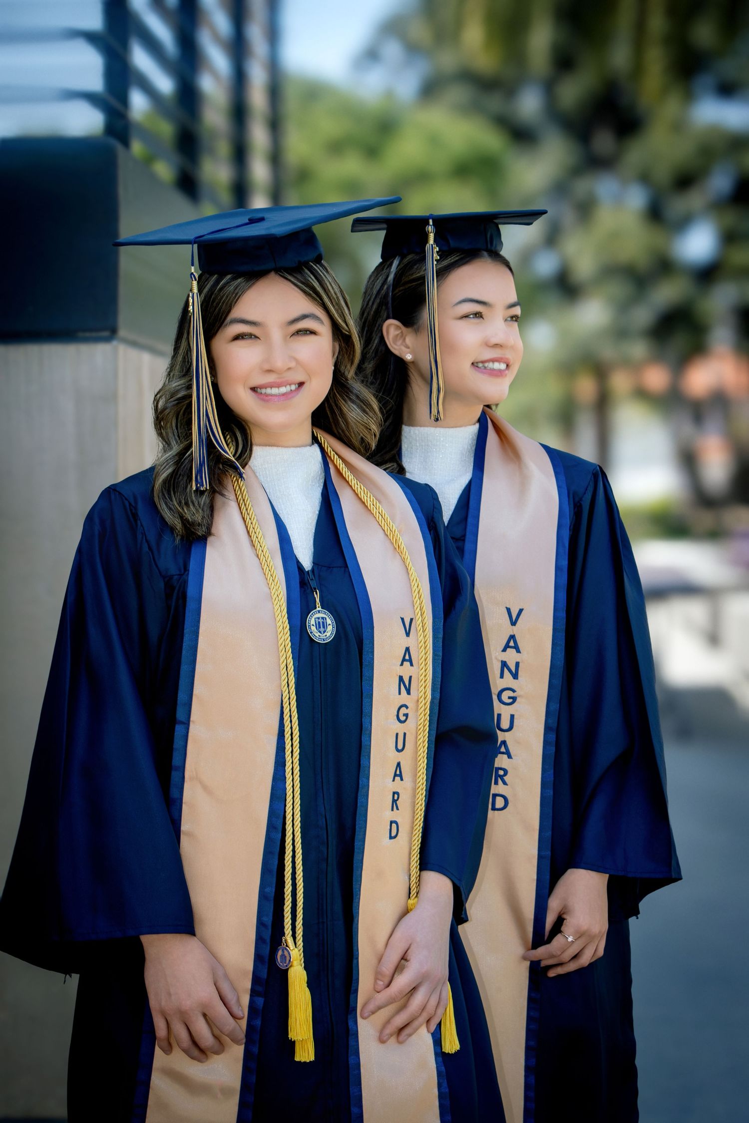 Two smiling women in navy Vanguard graduation gowns and caps pose proudly outdoors on a sunny day.