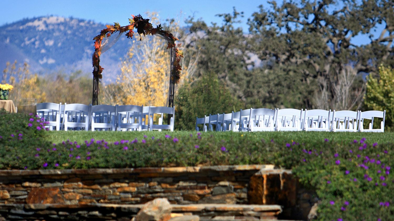 Outdoor wedding ceremony setup with floral arch, white chairs, purple wildflowers, and mountain backdrop under clear blue sky.