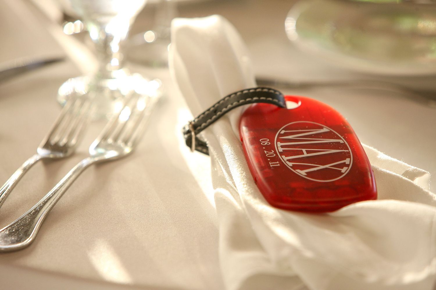 Elegant table setting with white napkin, silverware, and a red monogrammed luggage tag at a formal dinner.