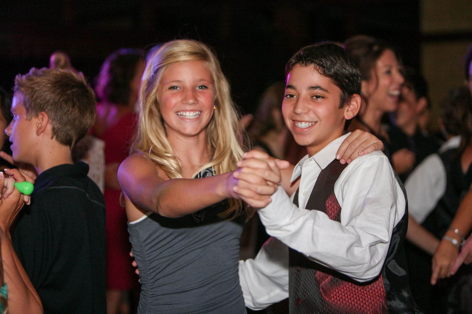 Two smiling teens dancing together at a formal party event, girl in gray dress and boy in white shirt with tie.