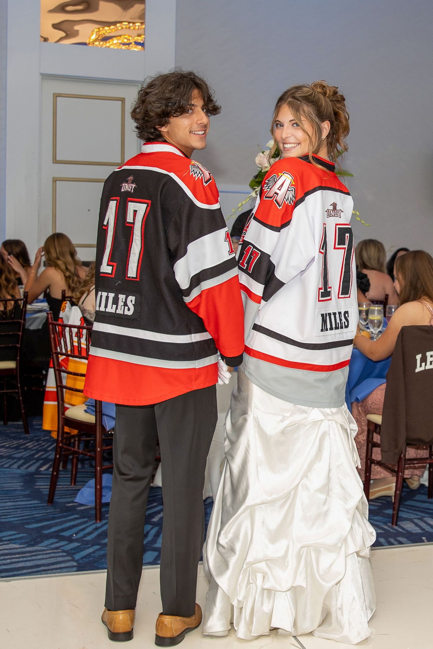 Couple in matching hockey jerseys number 17 posing back-to-back at prom, girl in white gown, boy in black pants.