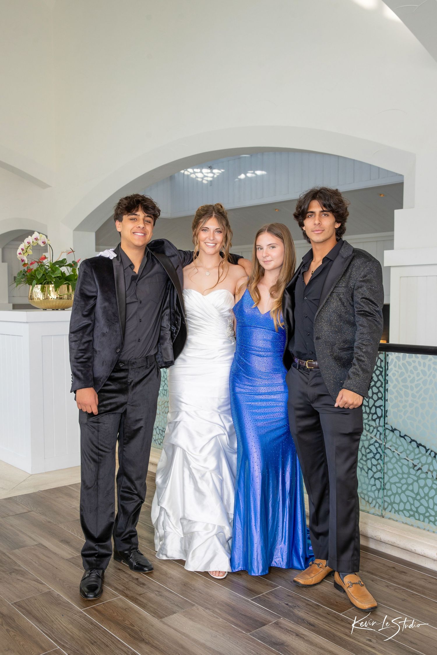 Four teens in formal attire, two in black suits and two in gowns, pose together at a prom event.