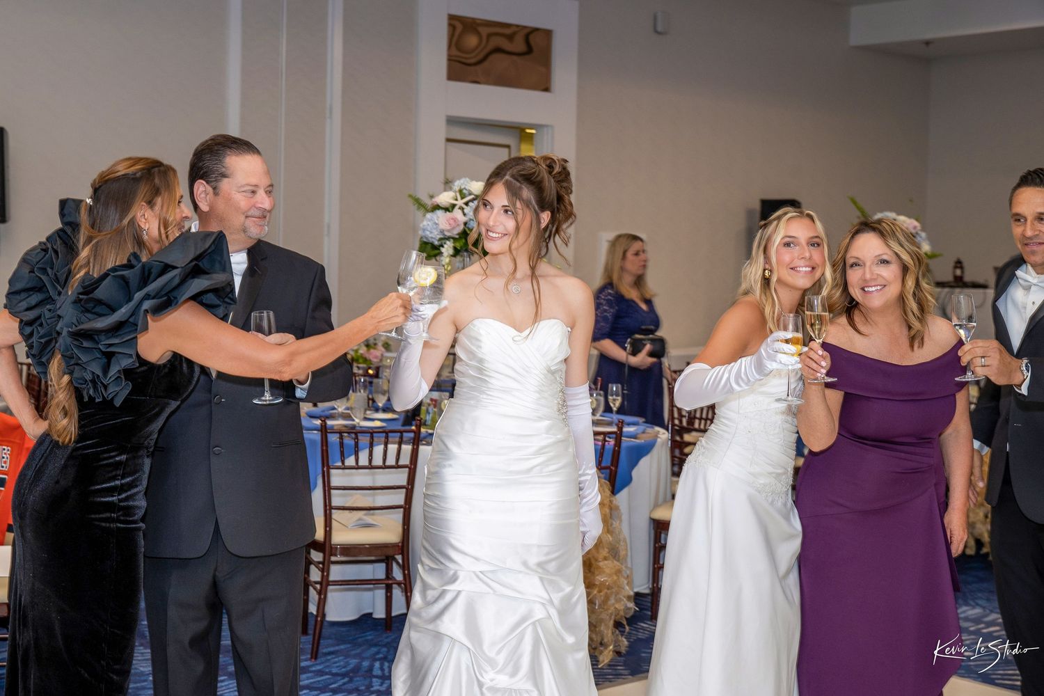 Bride in white gown toasting with wedding guests in elegant reception hall, bouquet raised in celebration.