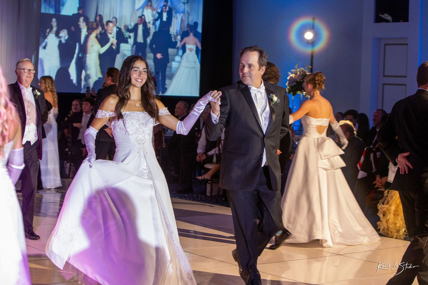 Elegant debutante ball with young woman in white gown dancing with man in tuxedo at formal event.