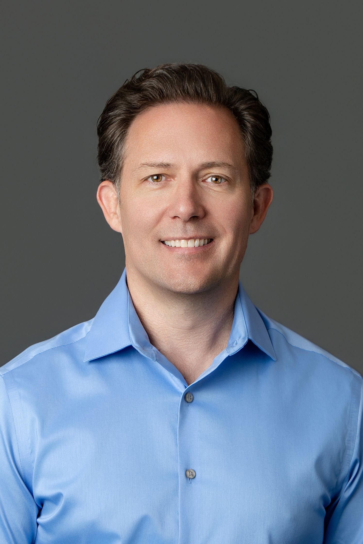 Professional headshot of a smiling middle-aged man with dark hair wearing a light blue button-up shirt against a gray background.