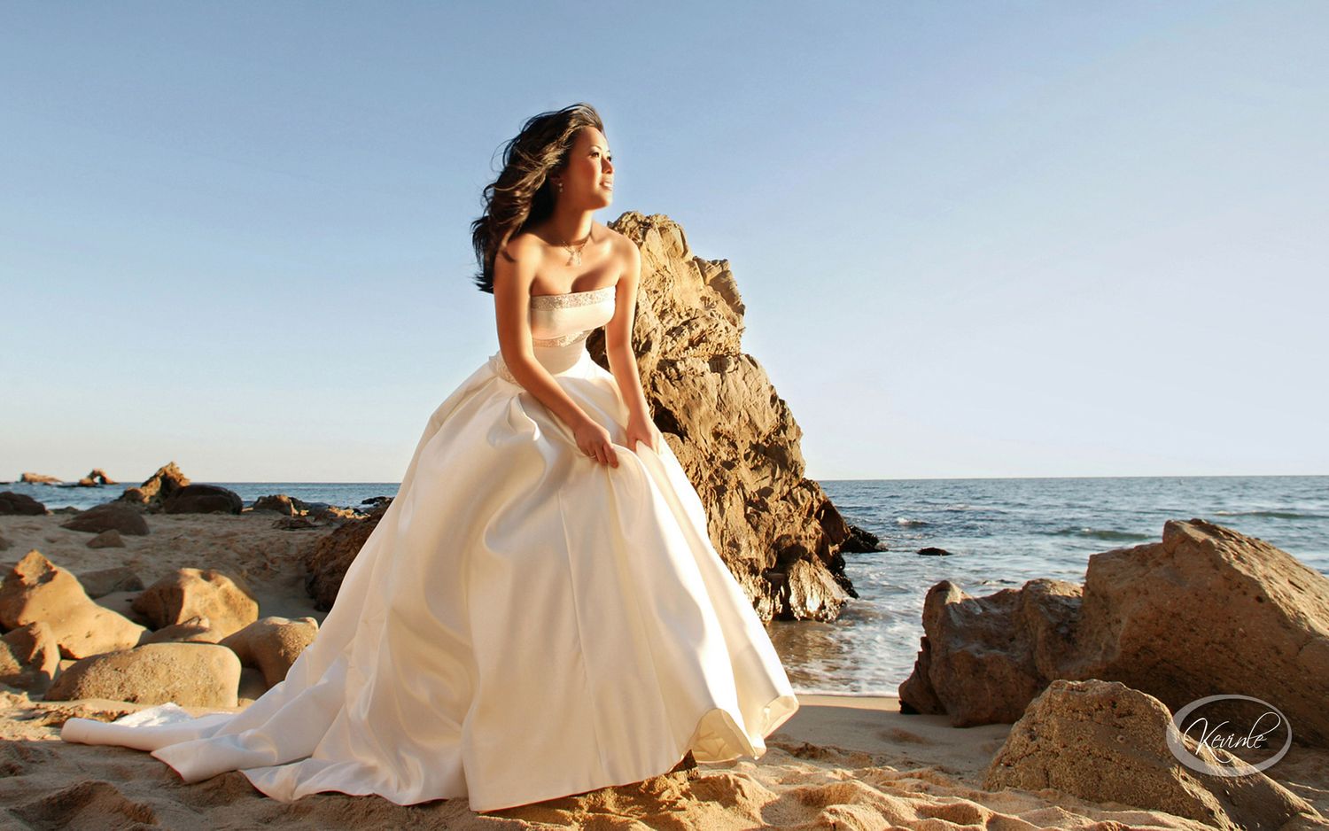 Bride in elegant white ball gown sitting on rocks by the ocean on a sunny beach day.