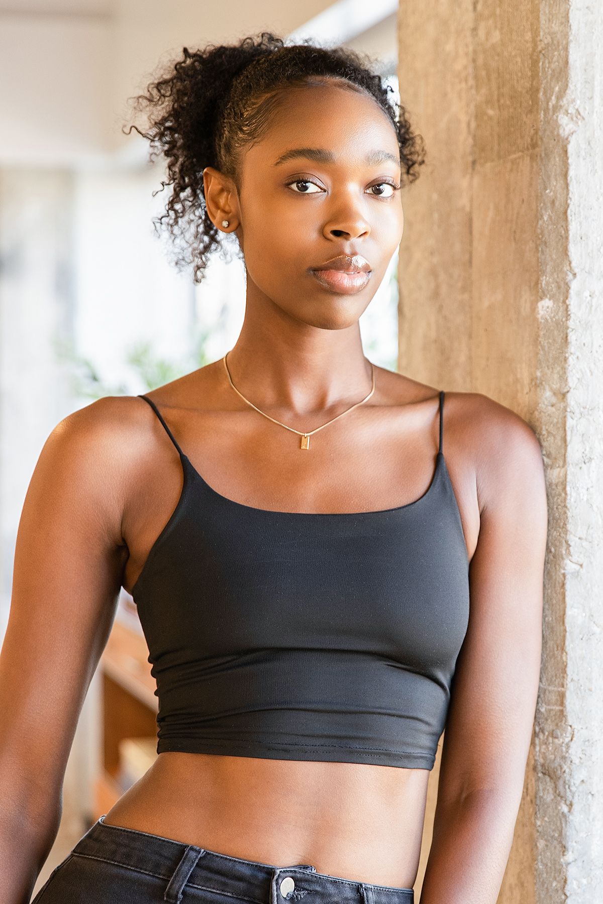 Young Black woman with curly ponytail wearing a black crop top and jeans with a gold necklace, leaning on a pillar.