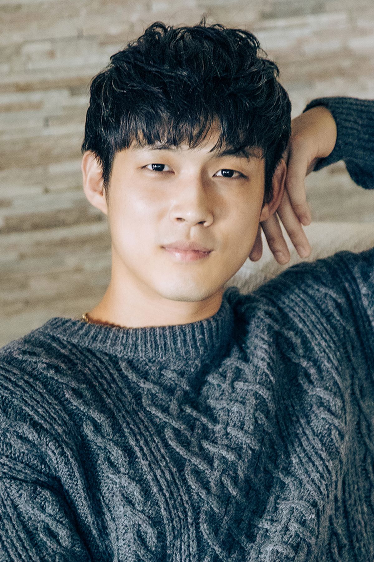 Young Asian man with dark wavy hair wearing a gray cable-knit sweater, posing with hand behind head.