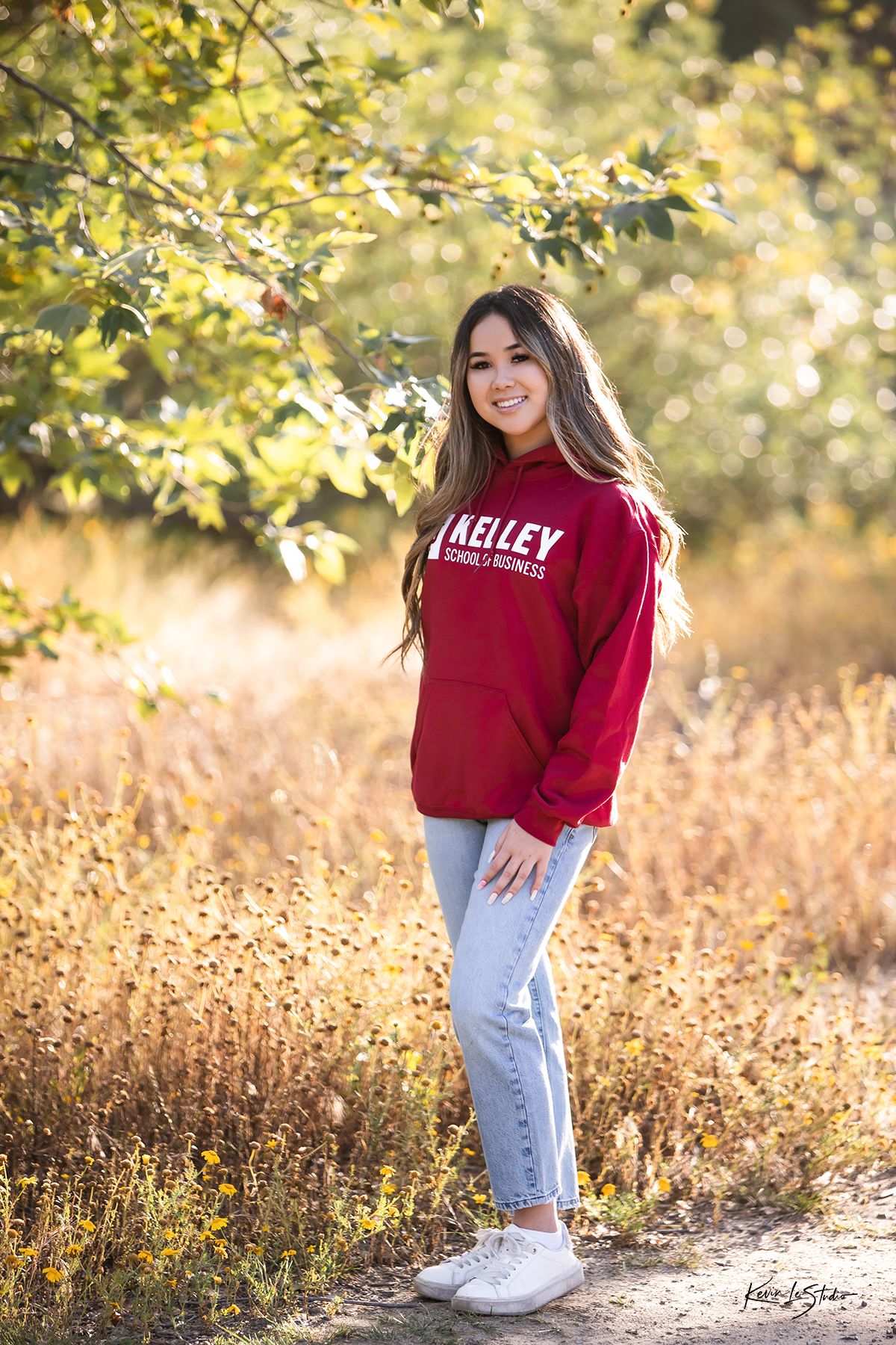 Smiling young woman in red Kelley School of Business sweatshirt poses on sunlit autumn nature trail path.