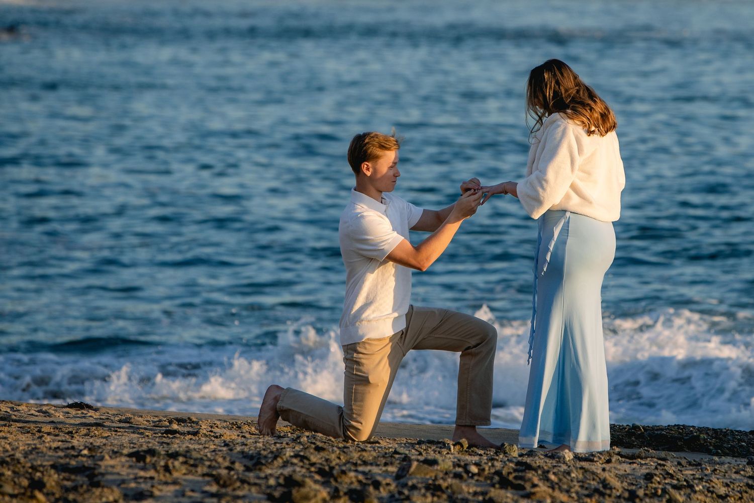 Man kneeling on beach proposing to woman in blue pants at sunset.