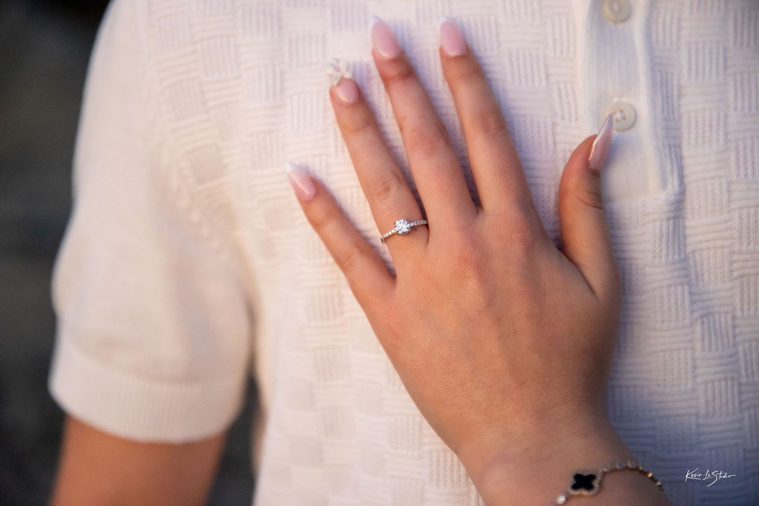 Close-up of a womans hand displaying a diamond engagement ring resting on a mans white knit shirt.