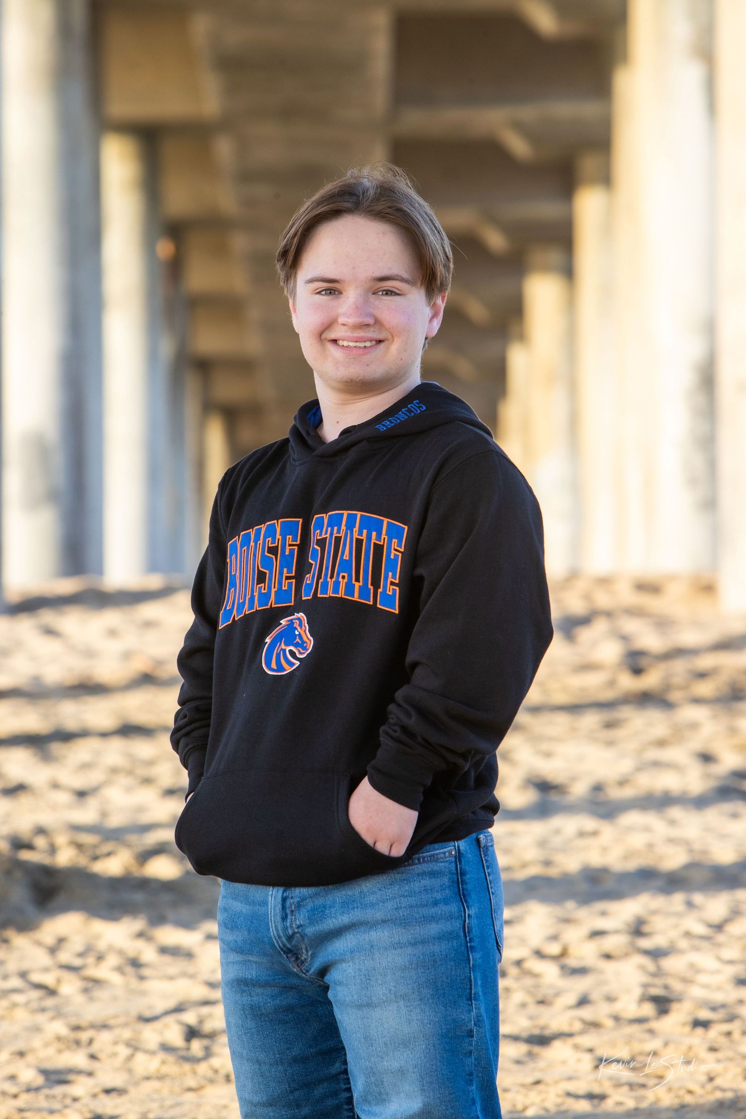 Teen smiling in Boise State hoodie under beach pier in golden light.
