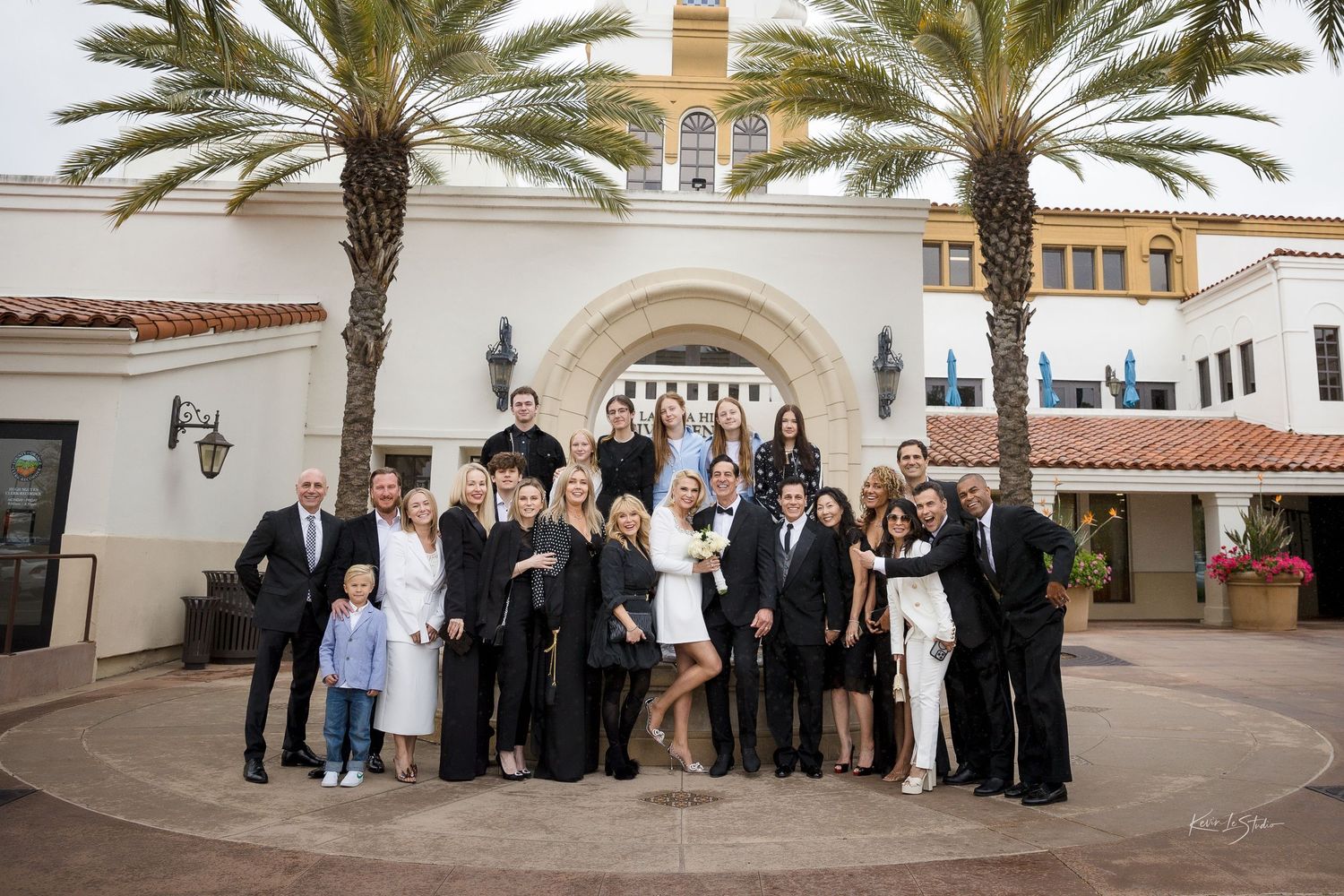 Wedding party poses in front of a Spanish-style venue with palm trees, guests dressed in black and white attire.