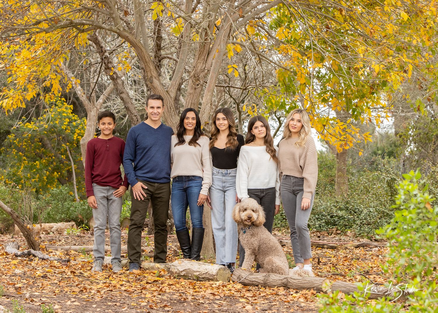 Family of five posing outdoors in autumn with golden leaves, trees, and a fluffy golden doodle dog sitting in front.