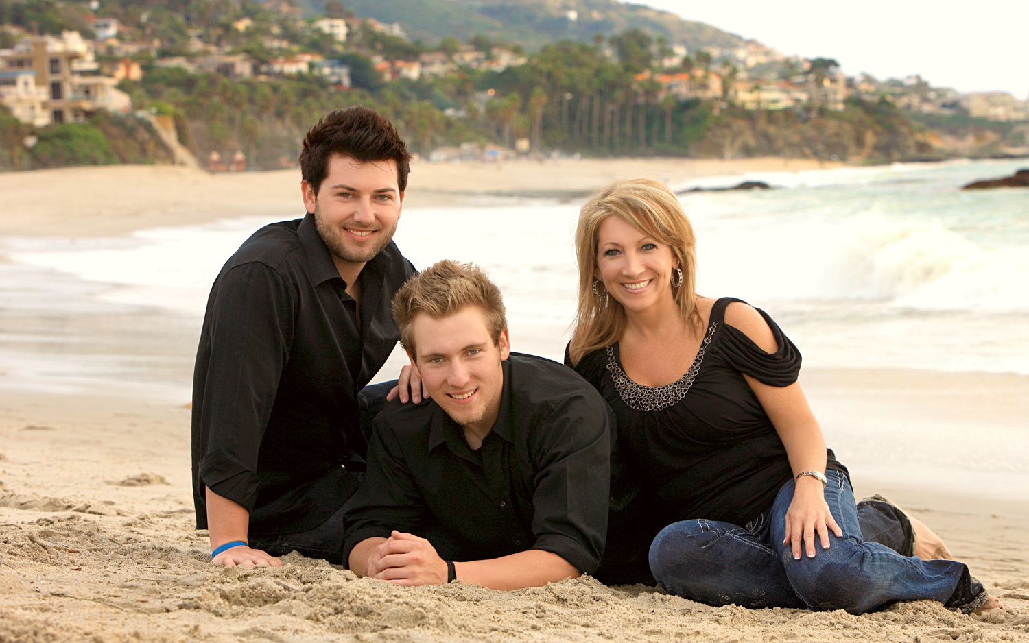 Family of three posing on a sandy beach, two men and a woman in black outfits, with ocean waves and cliffs in background.