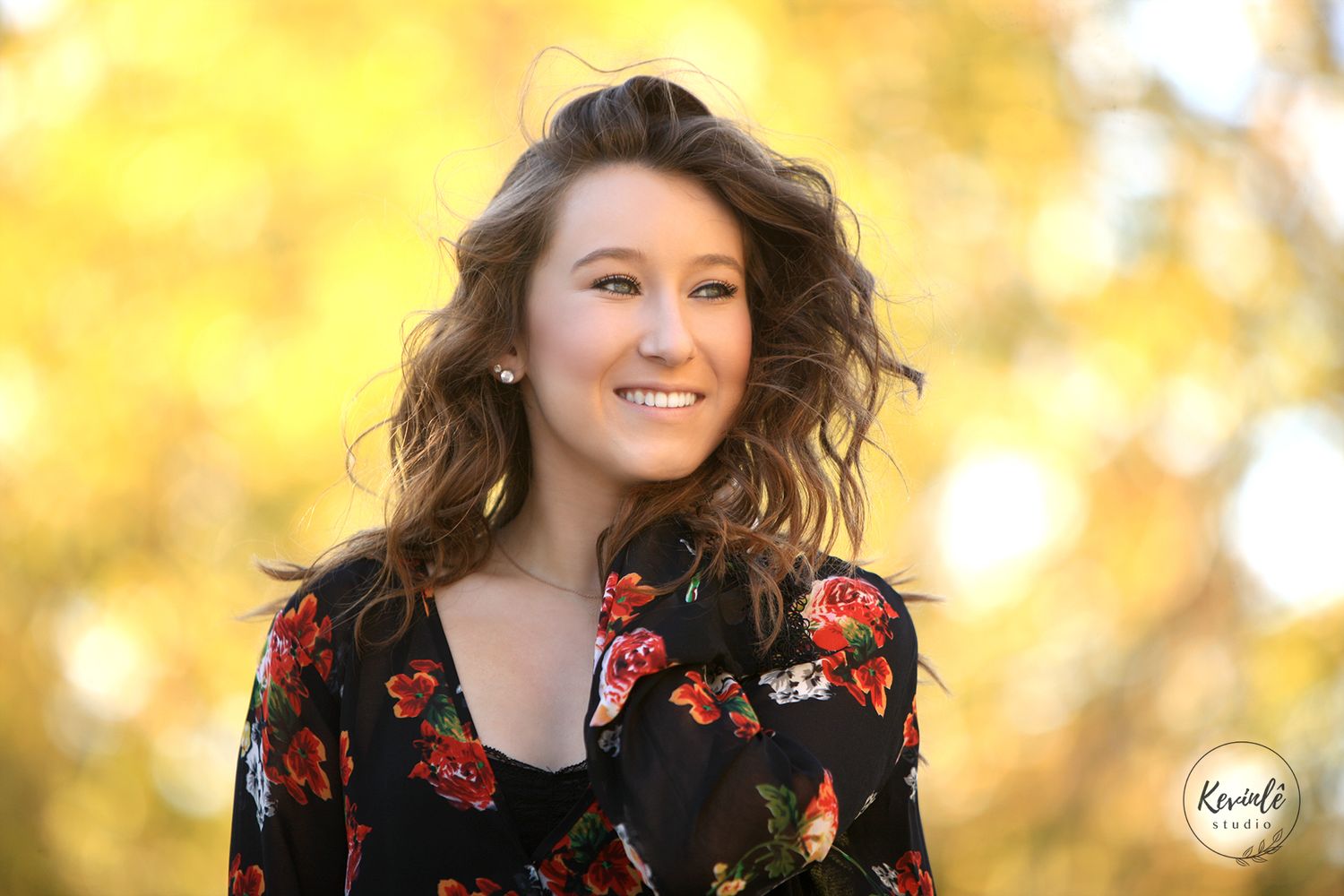 Young woman with curly brown hair smiling outdoors, wearing a floral dress, with golden autumn foliage background.