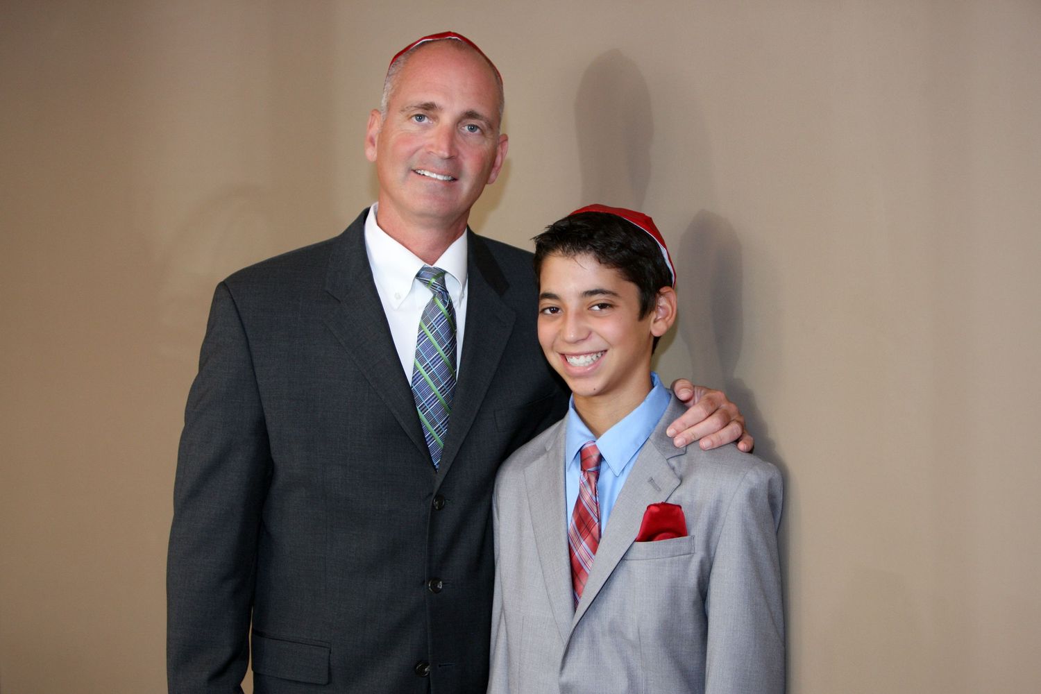 Two males in suits smiling for a formal photo, adult with arm around younger boy wearing red yarmulke.