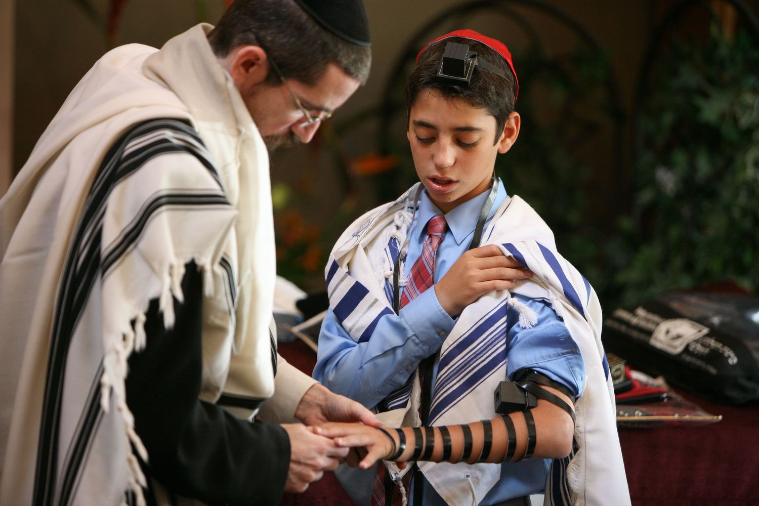 Rabbi assists a young boy during a Bar Mitzvah ceremony, both wearing traditional Jewish prayer shawls.