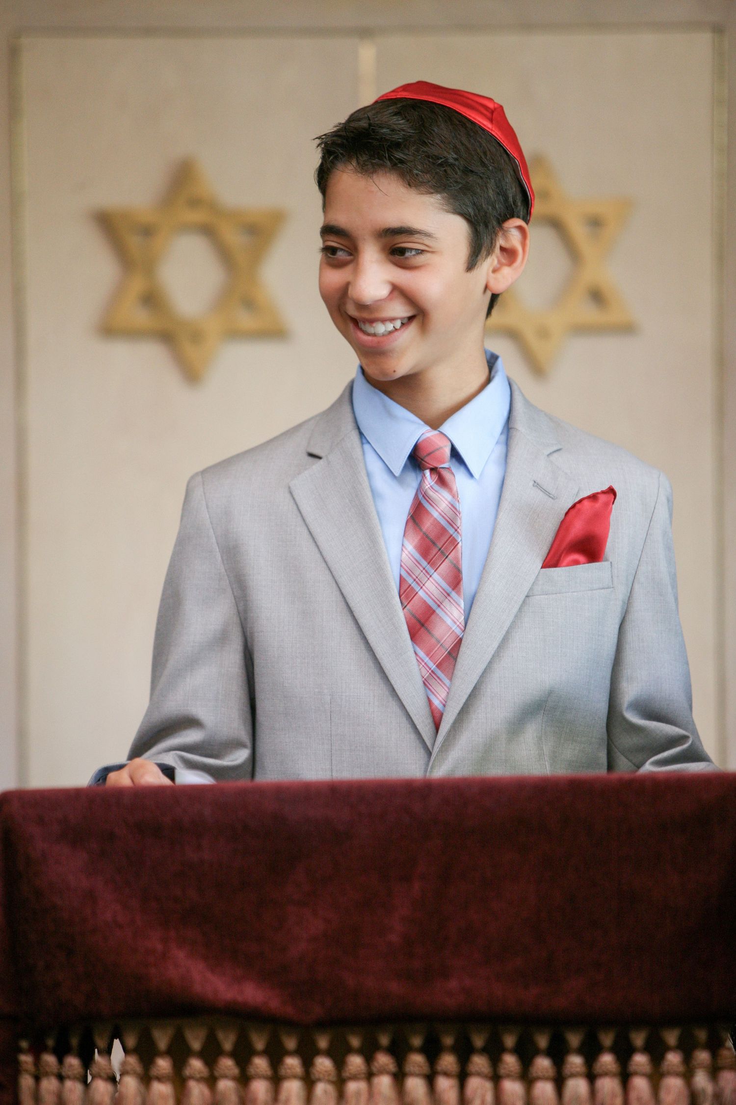 Smiling young boy in gray suit with red kippah stands at podium during Bar Mitzvah ceremony.