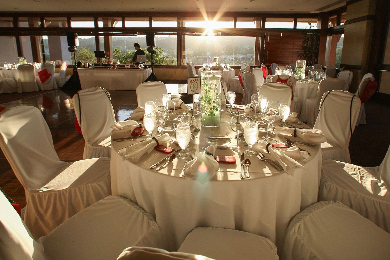 Elegant wedding reception venue with round tables, white chair covers, red accents, candles, and sunlit windows in background.