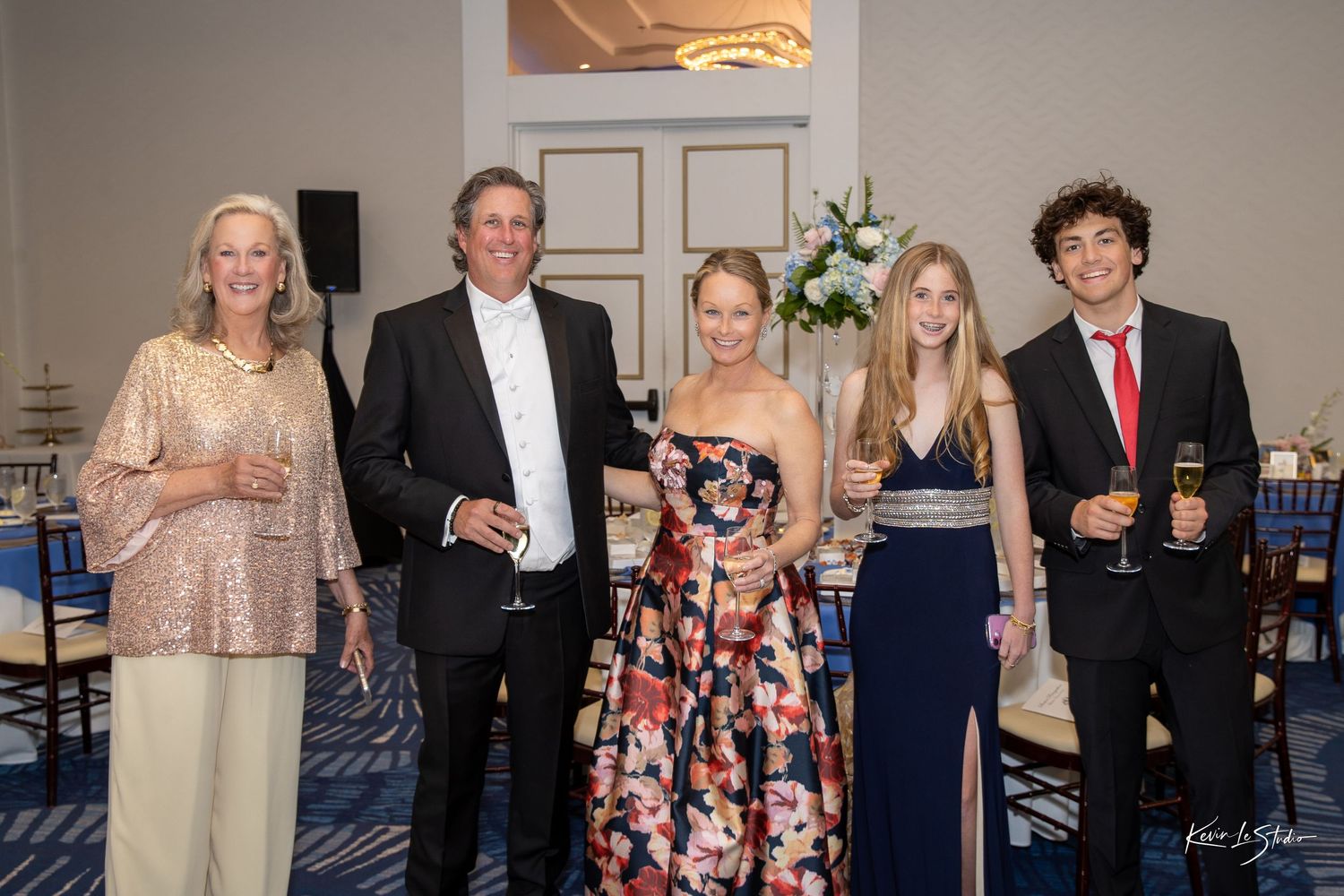 Five elegantly dressed guests pose together at a formal gala event in a decorated ballroom.
